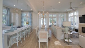 A spacious kitchen with white cabinetry, a dining area with white chairs, and a living room with a sofa and a TV. - Last minute discount July 5-11!  Watercolor Camp district 5BR w/ Pool & Spa (Santa Rosa Beach)