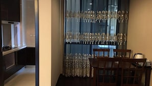 A dining area with a table and chairs, a window with decorative curtains, and a kitchen visible in the background. - Zixinju Aparthotel (Changsha)