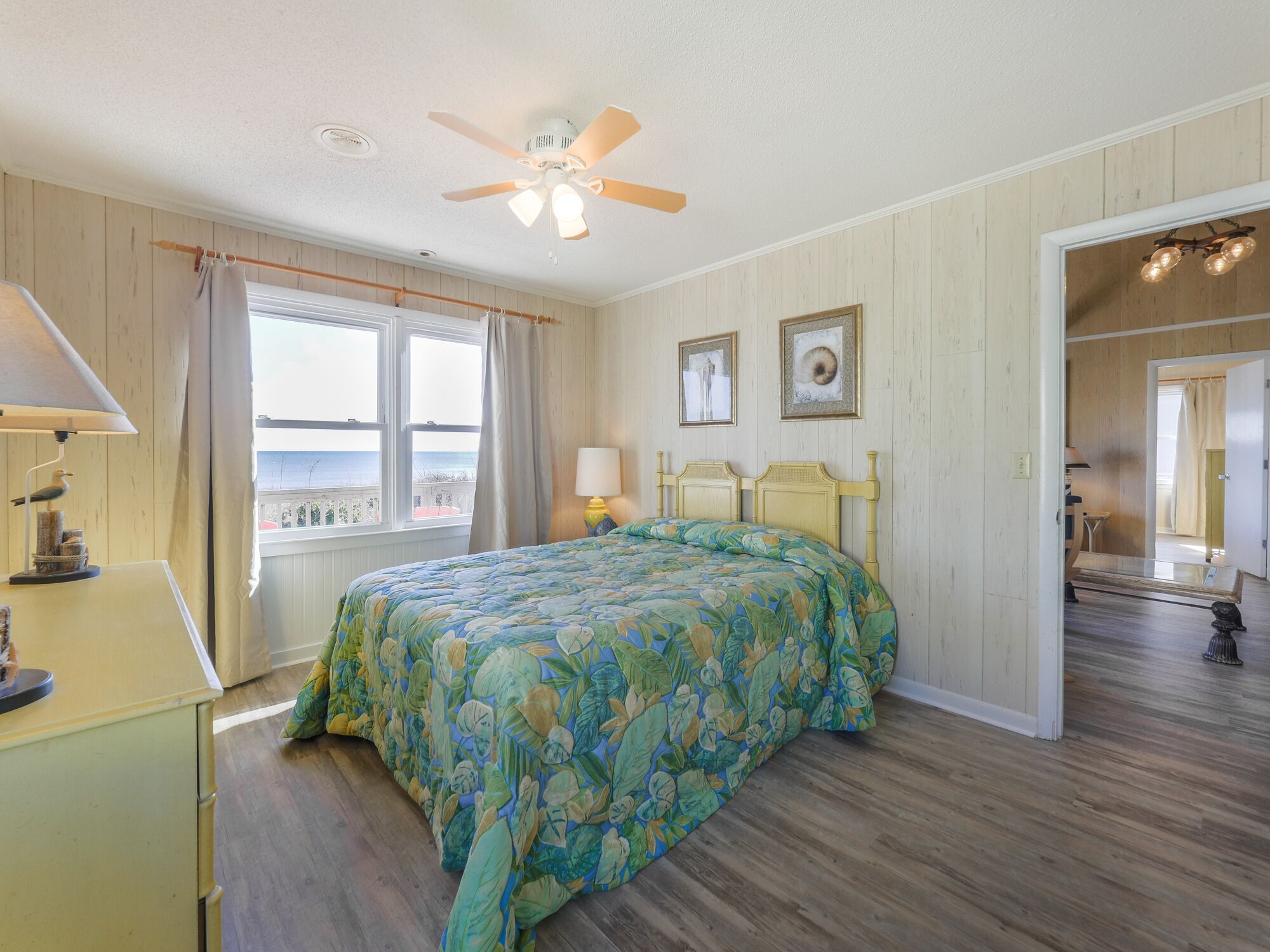 Une chambre à coucher comprenant un lit, un ventilateur de plafond, une fenêtre avec des rideaux, une commode et une porte donnant sur une autre pièce.