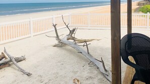 En vy över stranden från ett avskärmat område med ett vitt staket, ett trädäck och en svart stol.