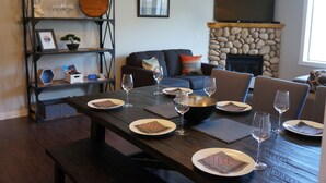 A dining area with a wooden table set for four, surrounded by chairs. A stone fireplace and a television are visible in the background.