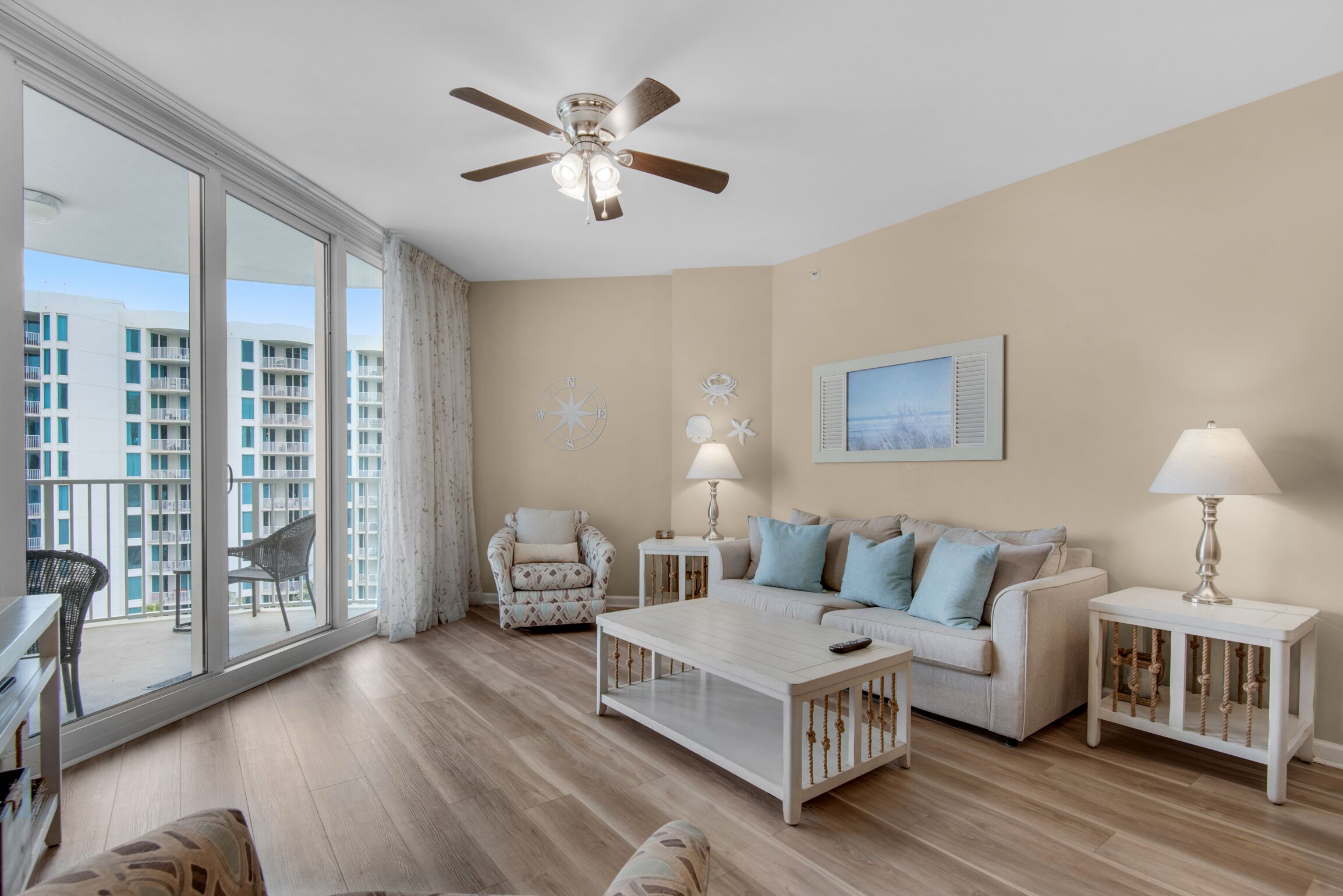 A spacious living room with a large window, a ceiling fan, a white coffee table, a beige sofa, and a patterned armchair.