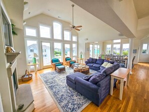 A spacious living room with a large sofa, a coffee table, and a fireplace. There are multiple windows providing natural light and a view of the outside.