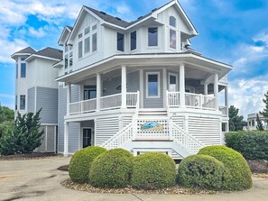 A two-story house with a balcony, a front porch, and a sign with a logo. - Ocean-side 6BR Corolla escape with private pool, hot tub & game room BI-20 (Corolla)