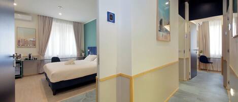 A hotel room with a bed, desk, and chair, a mirror, and a view of the cityscape through the window.