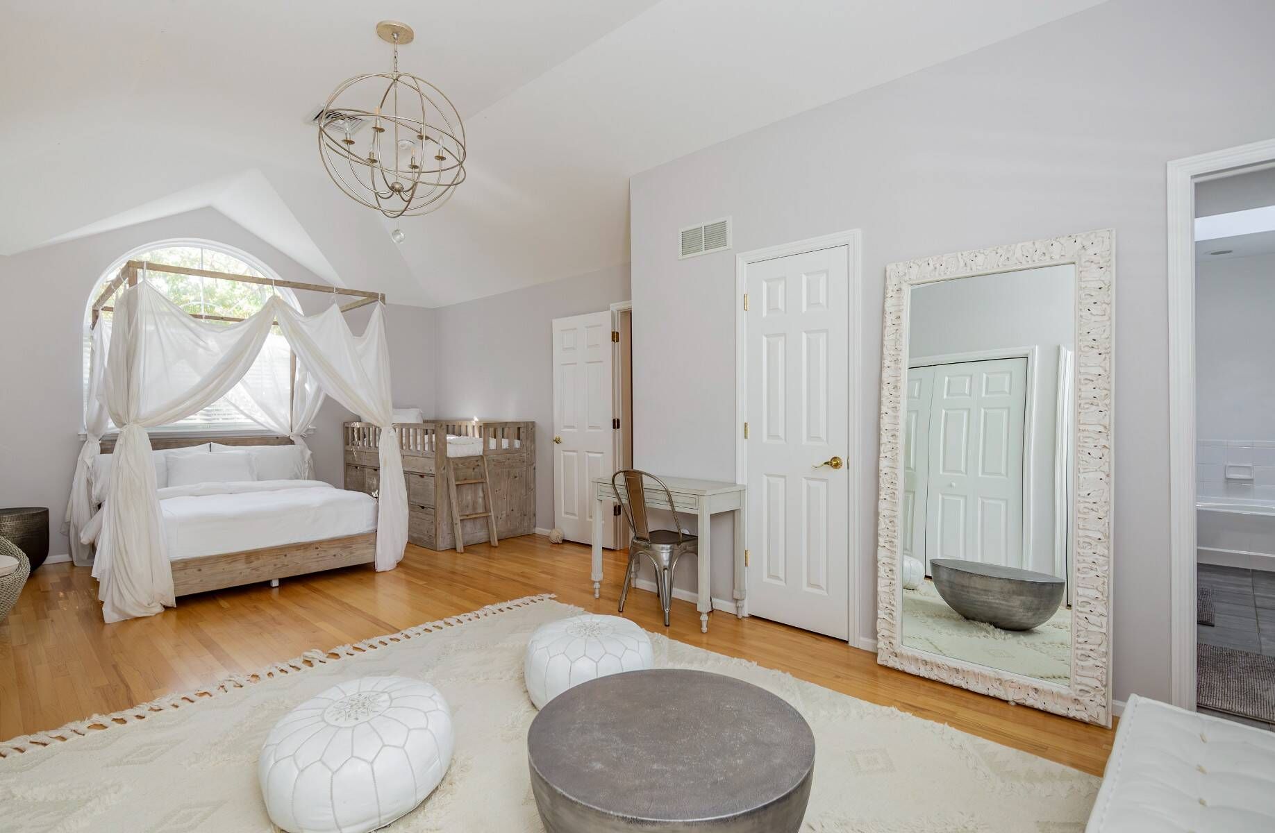 A bedroom with a canopy bed, a desk, a chair, a round ottoman, and a large mirror.