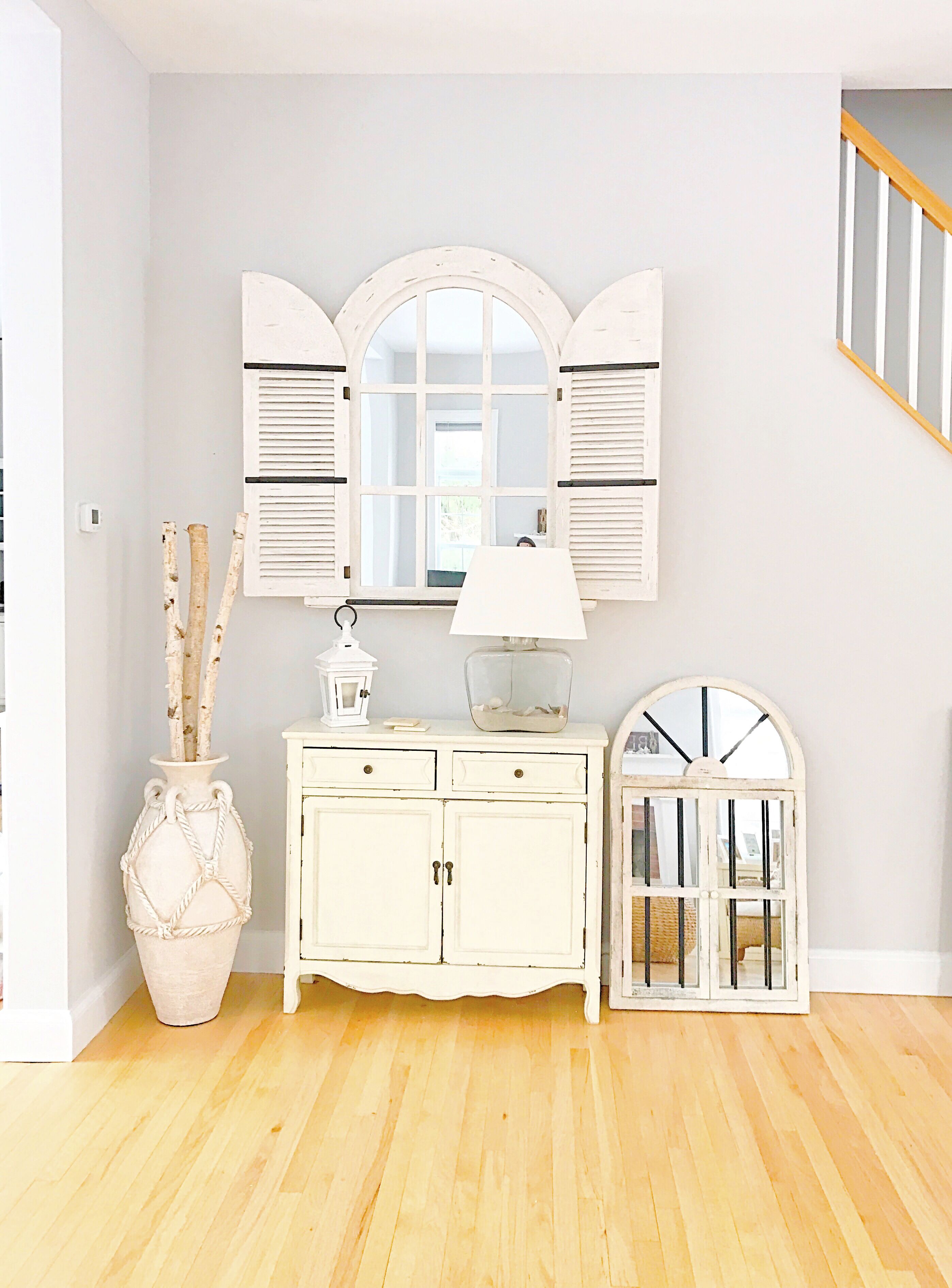 Une pièce avec une armoire blanche, un miroir à volets, un vase avec des branches et une petite table avec une lampe.