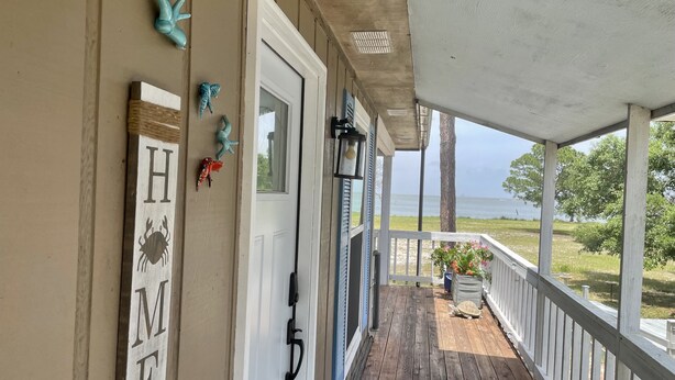 Une véranda en bois offrant une vue sur l’océan, une porte sur laquelle est inscrit le mot « MAISON », et des poissons décoratifs.