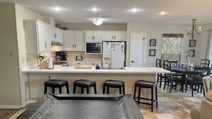 Une cuisine moderne avec des meubles blancs, un îlot central doté de tabourets, un coin repas avec une table et des chaises, et un lustre.