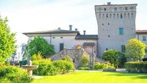 A historic building with a prominent tower, surrounded by well-maintained gardens and a clear blue sky. - La Tavola Rotonda (Cortemaggiore)