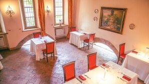 A dining area with tables set for four, each with red chairs. There are two windows with curtains, a painting on the wall, and a tiled floor. - La Tavola Rotonda (Cortemaggiore)