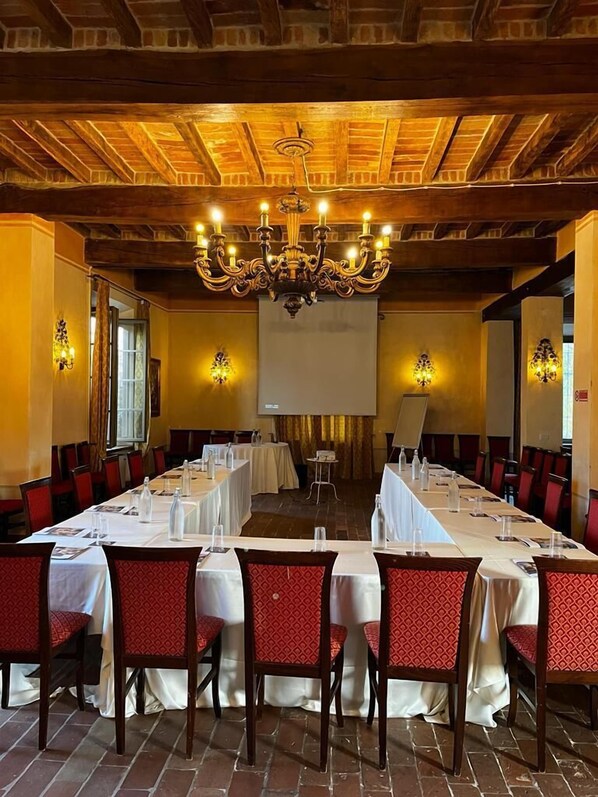 A banquet hall with long tables, red upholstered chairs, a chandelier, and a projector screen. - La Tavola Rotonda (Cortemaggiore)