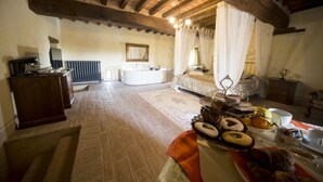 A rustic room with exposed wooden beams, a large bed with white curtains, a wooden dresser, and a table with pastries. - La Tavola Rotonda (Cortemaggiore)
