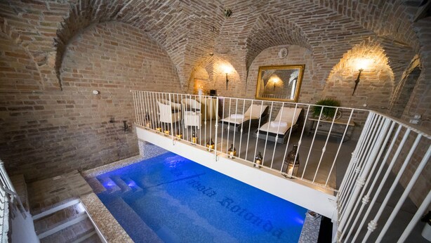 A swimming pool with a tiled edge and a white railing, surrounded by stone walls and a wooden staircase. - La Tavola Rotonda (Cortemaggiore)