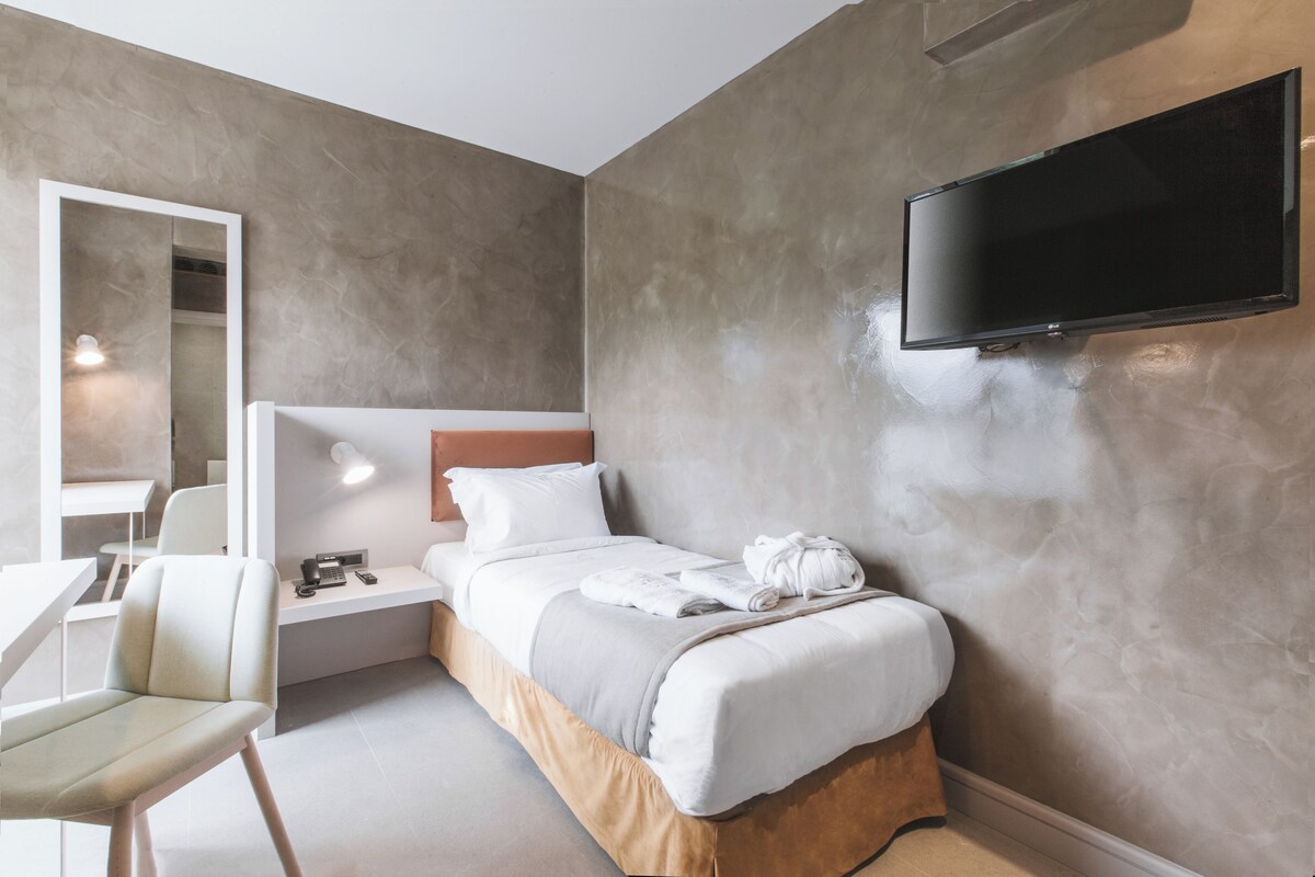A modern hotel room with a bed, a TV mounted on the wall, a desk with a chair, and a mirror.