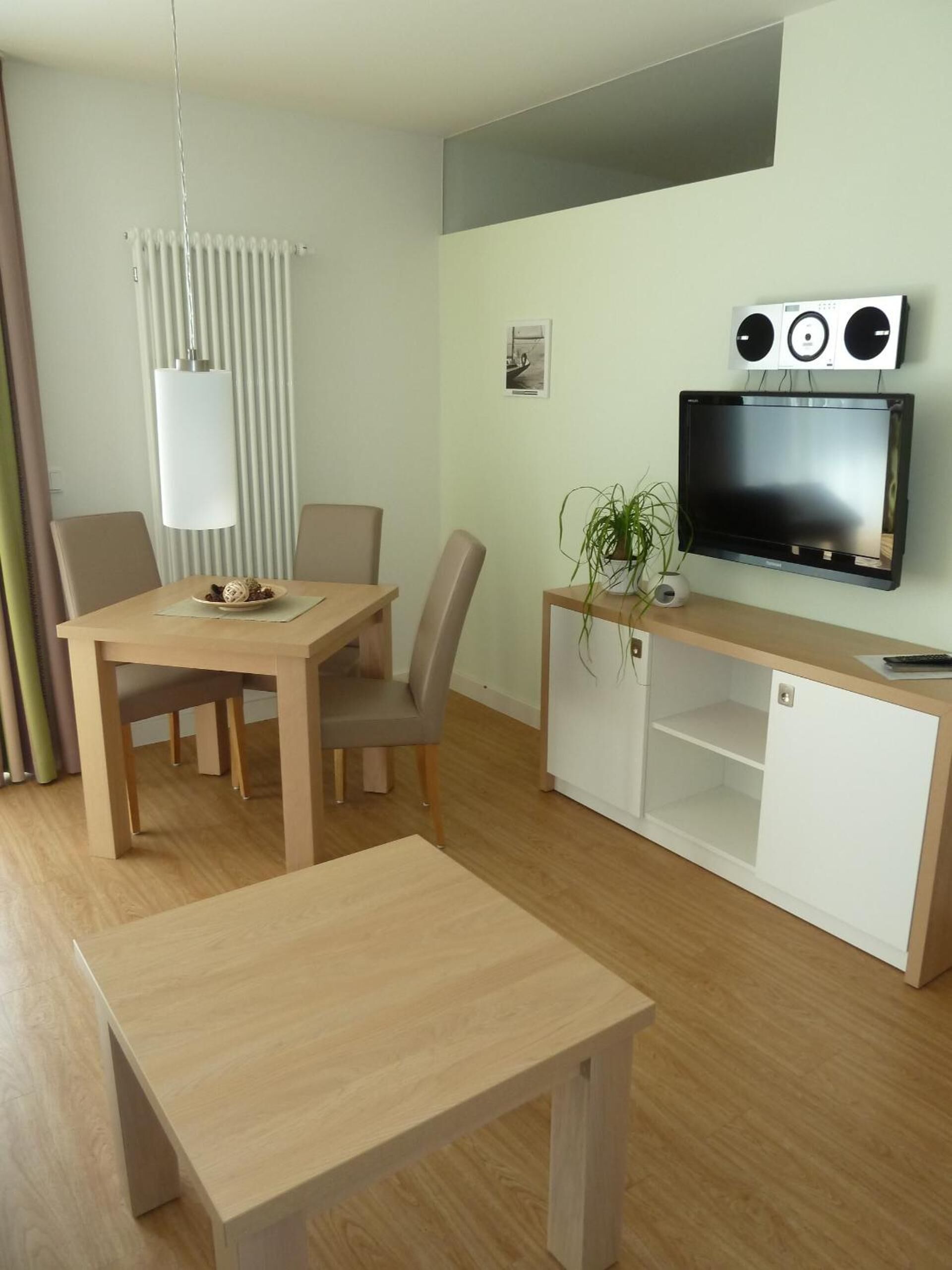 A modern living room with a wooden dining table, a TV mounted on the wall, and a small cabinet.
