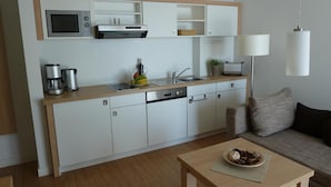 Uma cozinha compacta com micro-ondas, cafeteira e torradeira. Possui uma área de refeições com mesa e sofá.