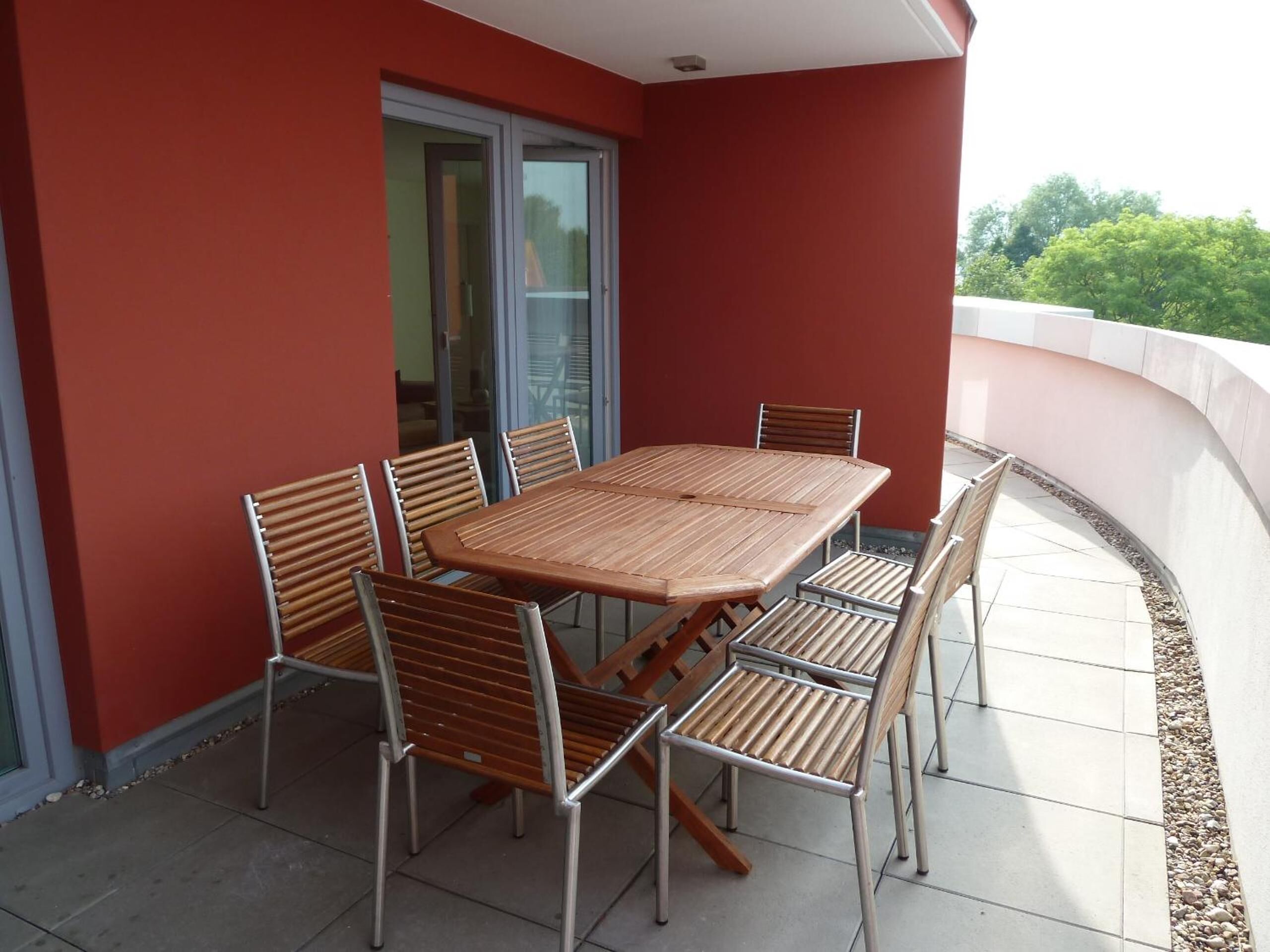 Un patio exterior con una mesa de madera y seis sillas, una puerta de cristal y una pared roja.