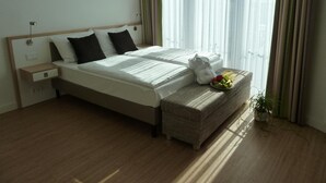 Quarto de hotel com uma cama grande, uma mesinha de cabeceira, um pequeno banco com frutas e uma planta em vaso.