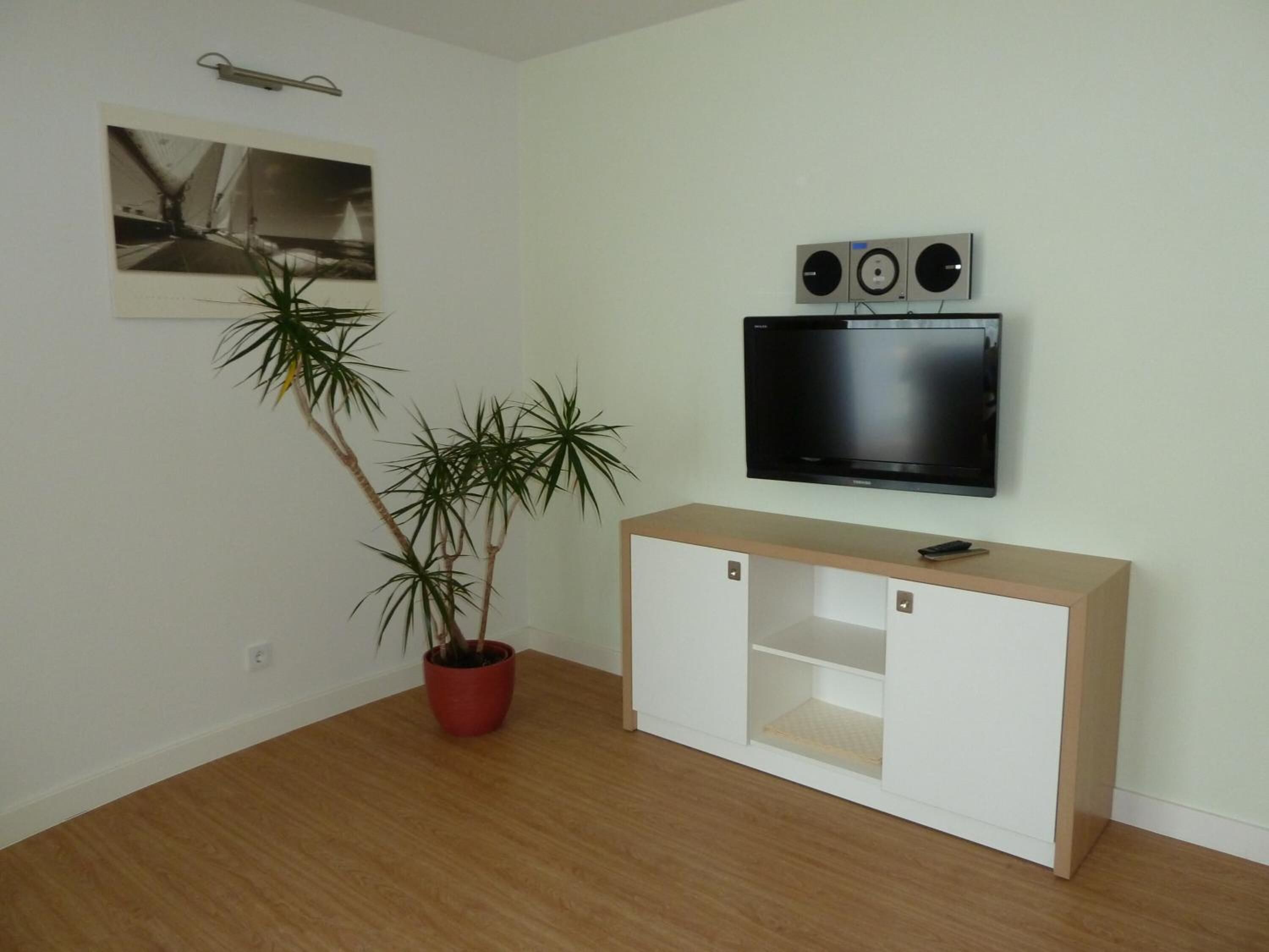 Una habitación con piso de madera, un mueble blanco con televisor y bocinas instaladas en la pared, y una planta en maceta.