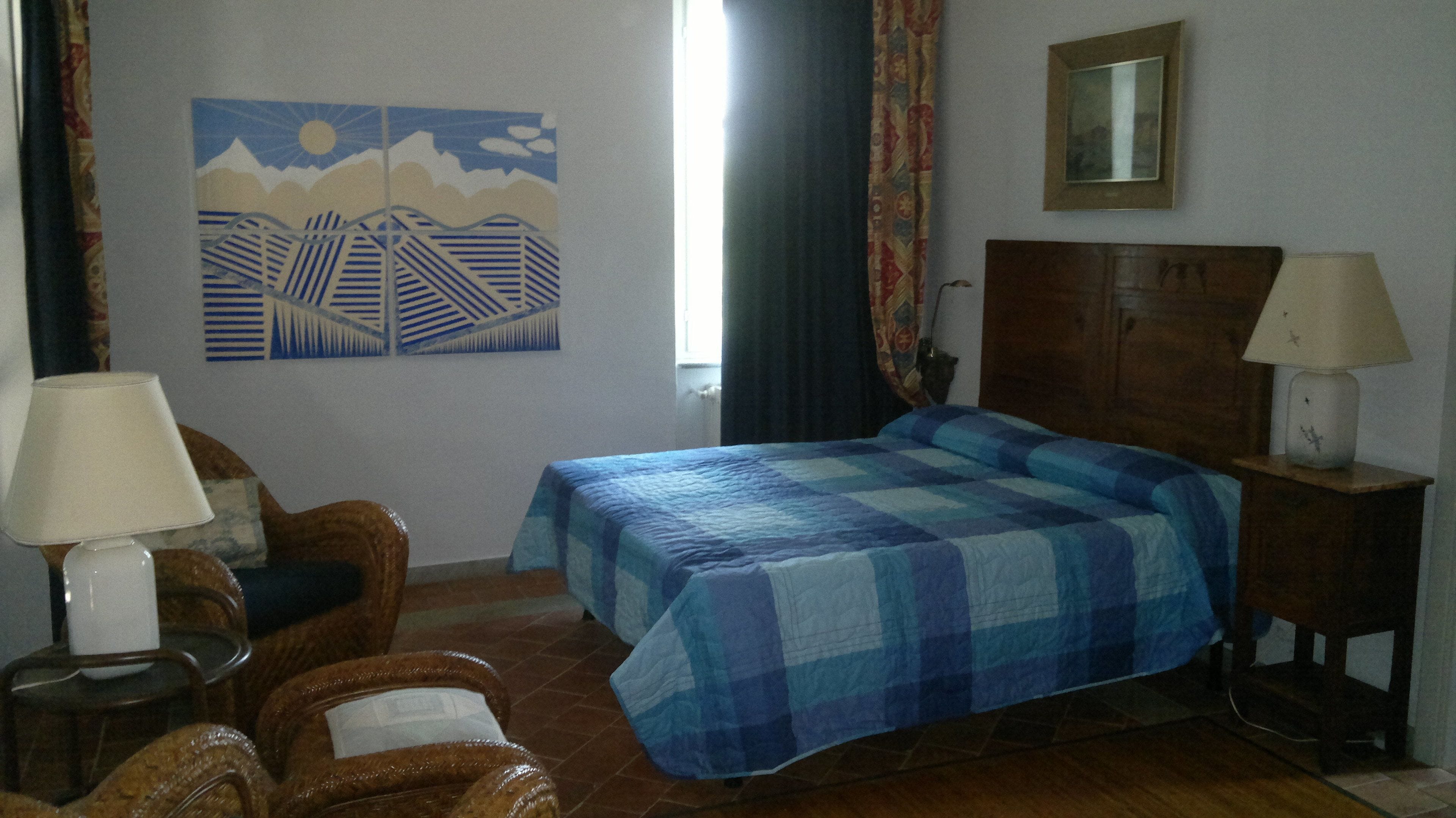A room with a bed, two chairs, a nightstand with a lamp, a painting on the wall, and a window with curtains.