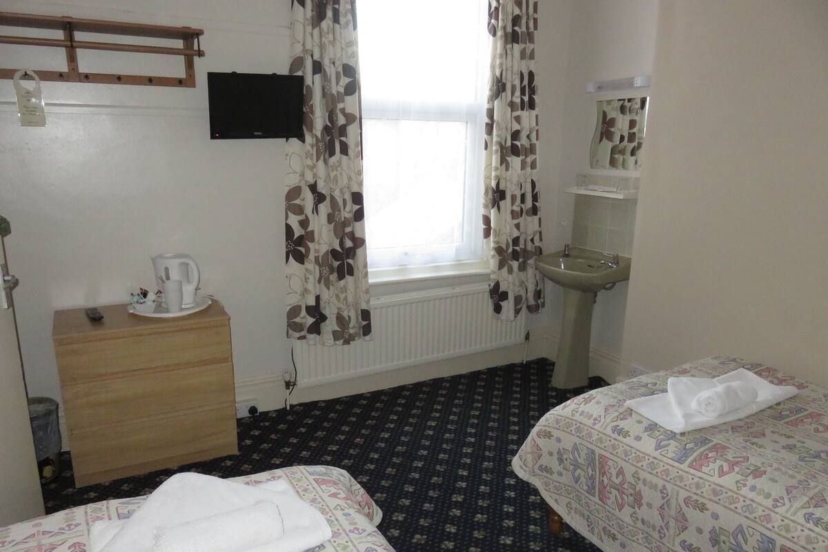 A hotel room with two beds, a wooden dresser, a television, a window with curtains, a sink, and a radiator.