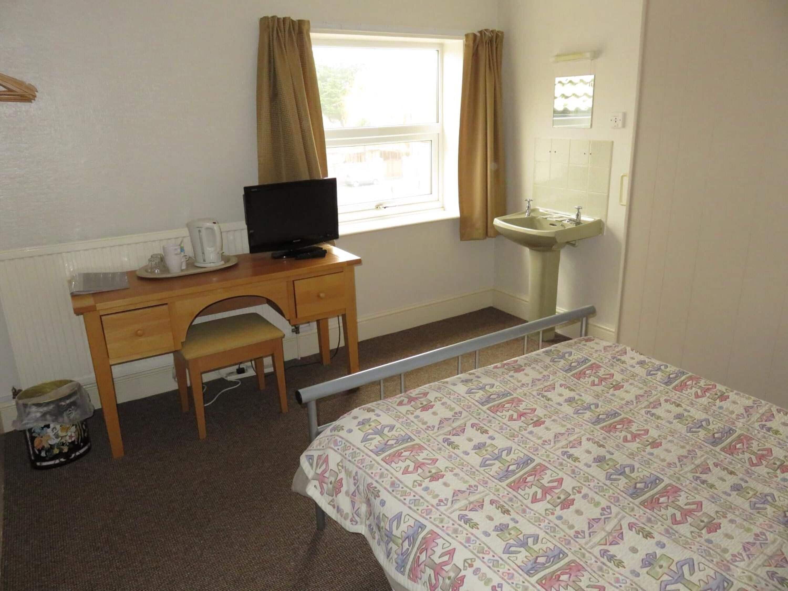 A bedroom with a bed, a desk with a television, a chair, a window with curtains, a sink, and a mirror.
