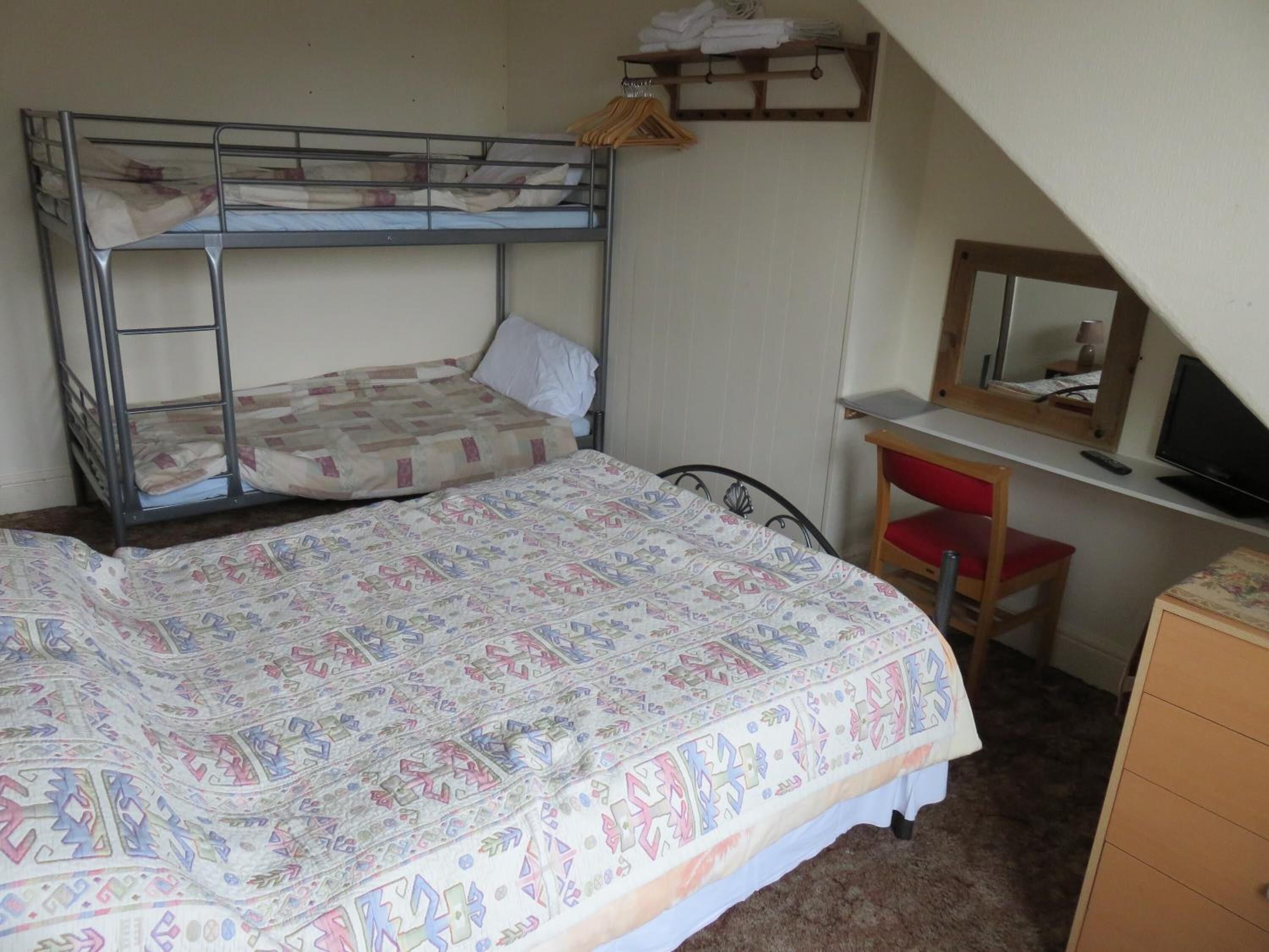 A dormitory room with bunk beds, a desk with a chair, a mirror, and a dresser.