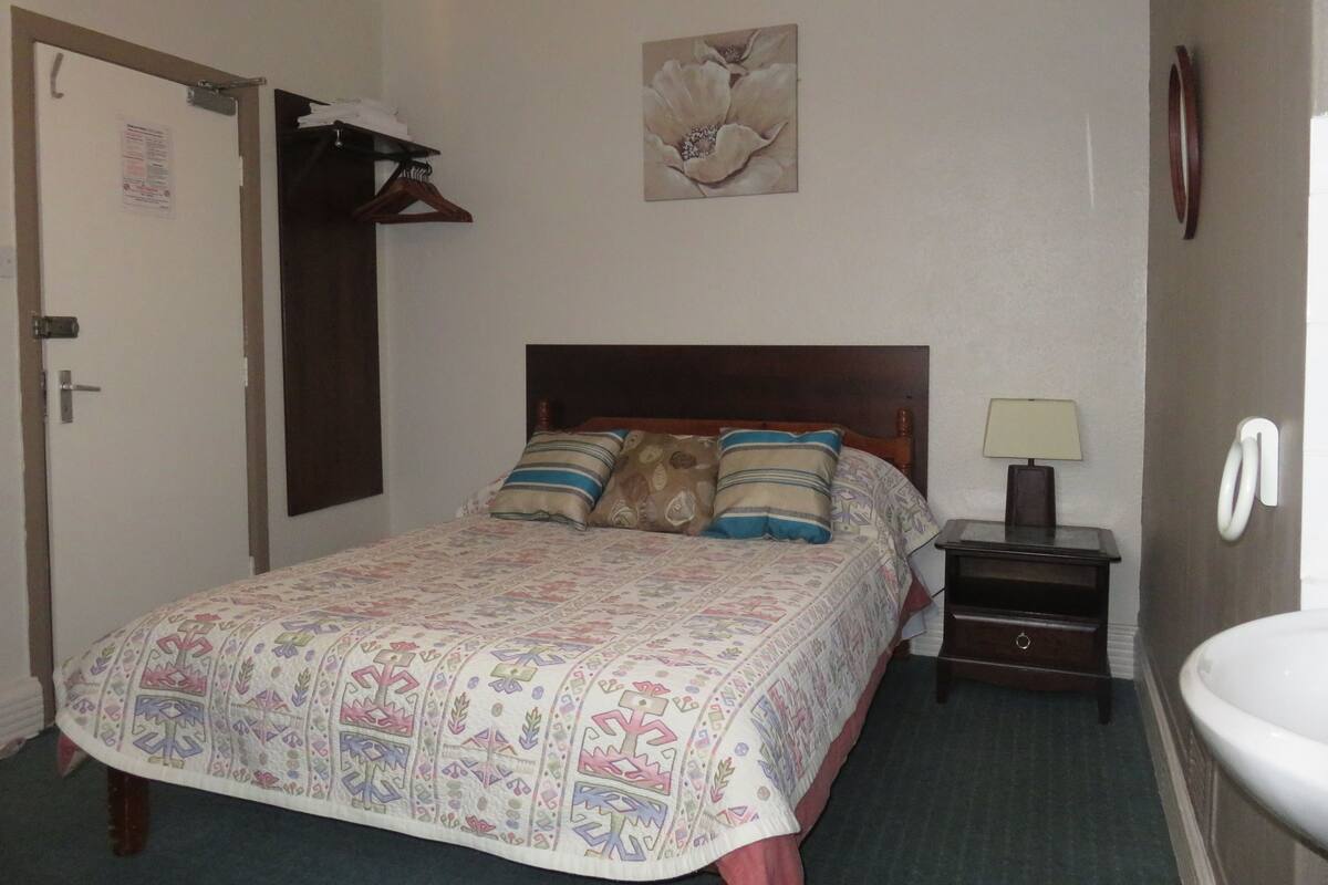 A neatly made bed with patterned bedding, a bedside table with a lamp, and a wall-mounted toilet.