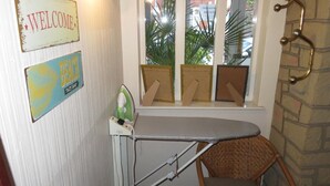 A corner with a wall-mounted ironing board, a wicker chair, and a window with a view of palm trees. - Glencoe Hotel (Bridlington)