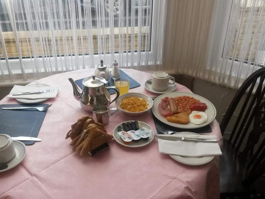 A breakfast table with a pink tablecloth, plates of food, a teapot, cups, and a chair.