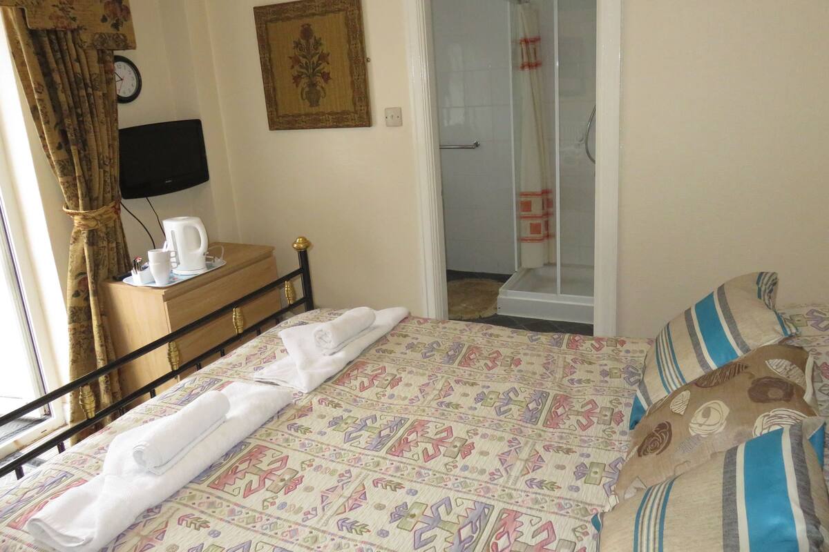 A hotel room with a bed, a bedside table with a kettle, a clock, and a shower area.