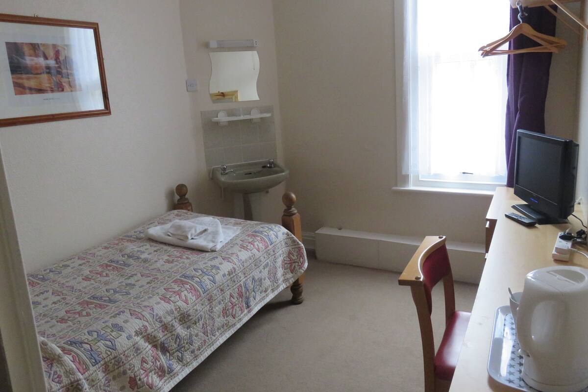A small, simple room with a bed, a desk, a chair, a sink, and a window.