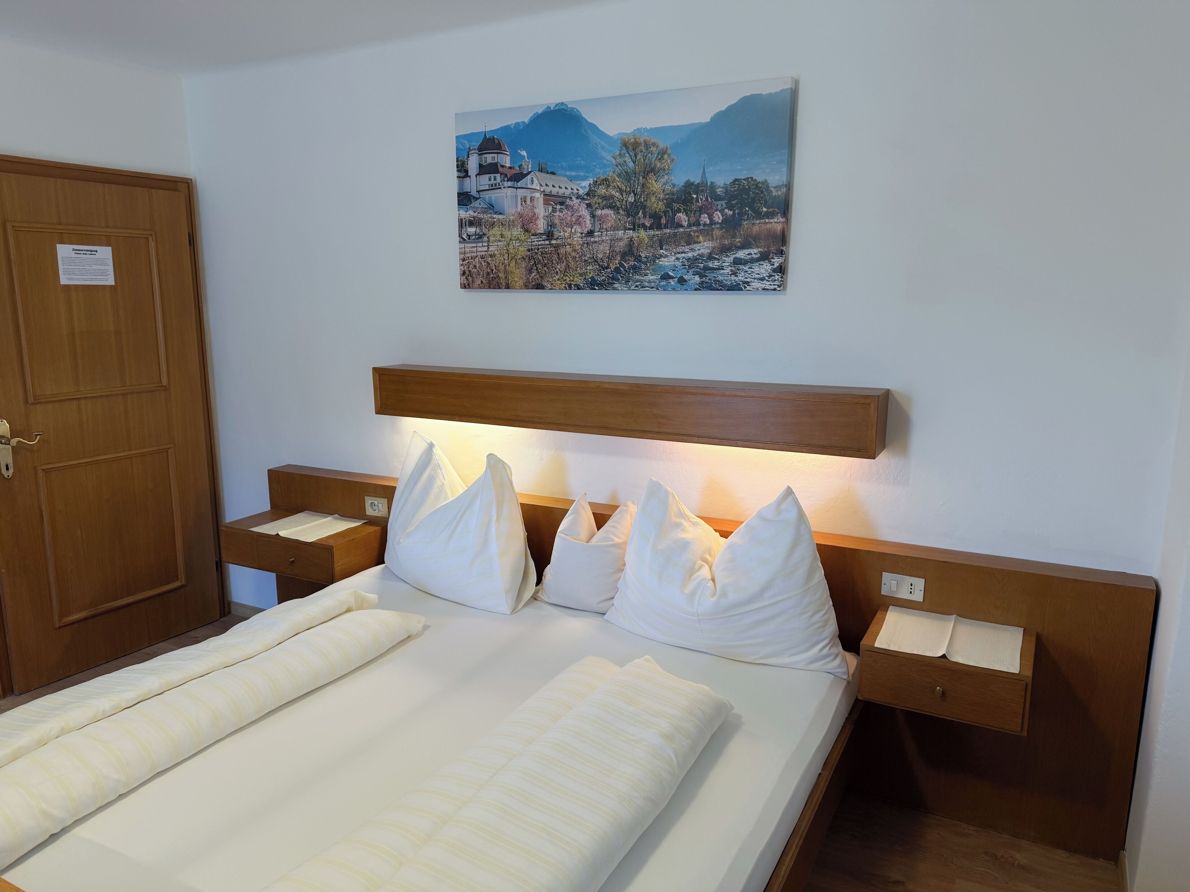 Een hotelkamer met een bed, een houten hoofdbord, witte kussens, een nachtkastje en een schilderij van een berglandschap.