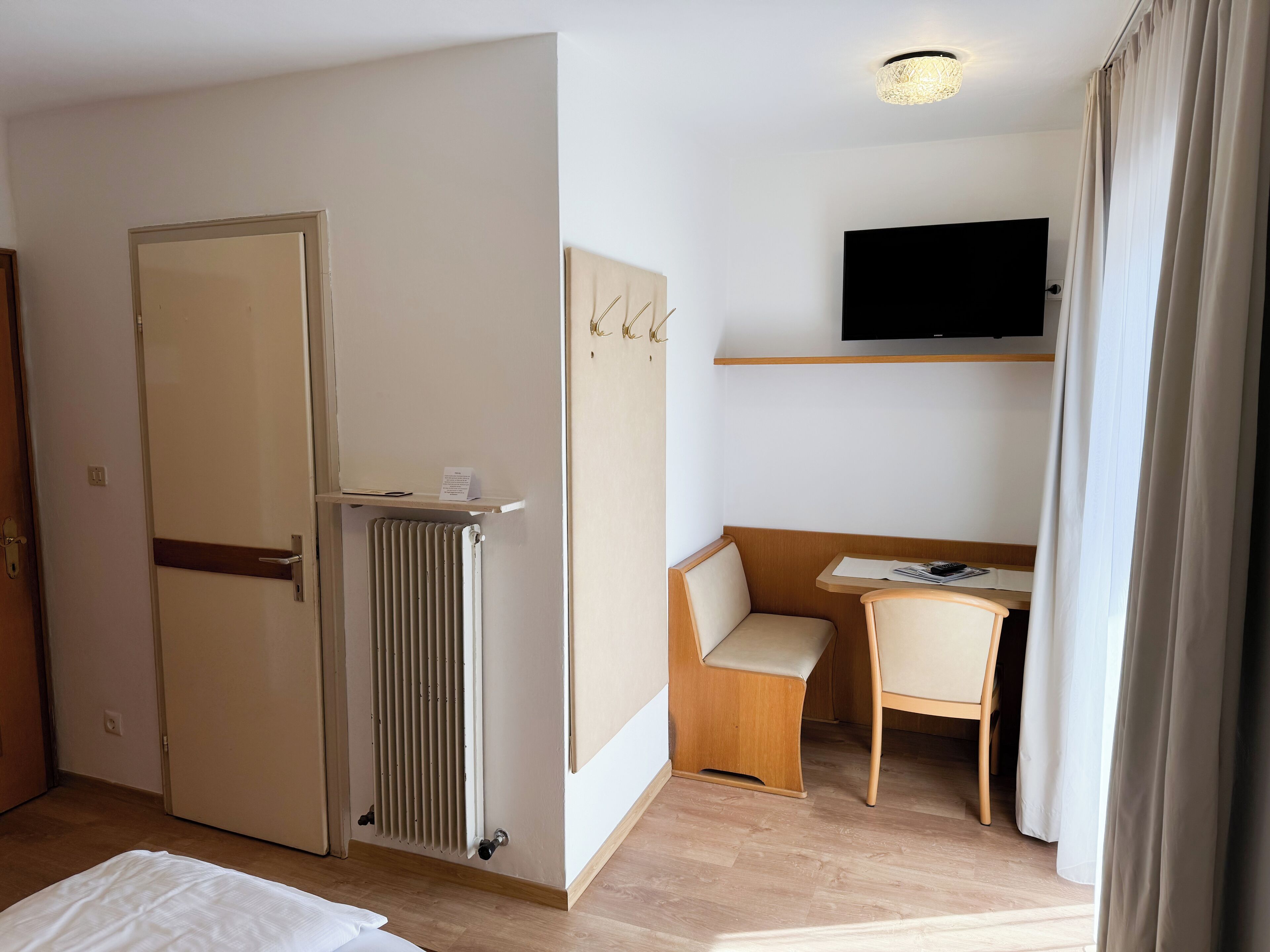 Una camera d'albergo con un letto, una scrivania con una sedia, un termosifone, una televisione fissata al muro e una porta.