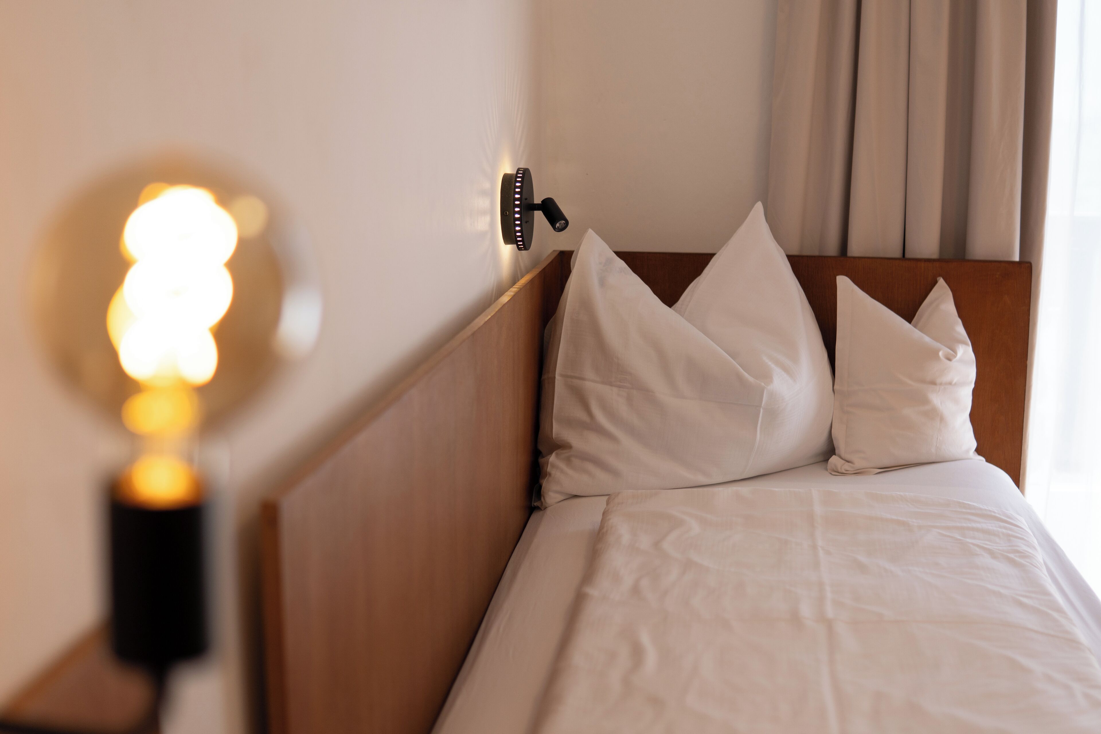 Ein ordentlich bezogenes Bett mit weißen Leinen und Kissen, ein Holz-Kopfteil und eine Wandleuchte.