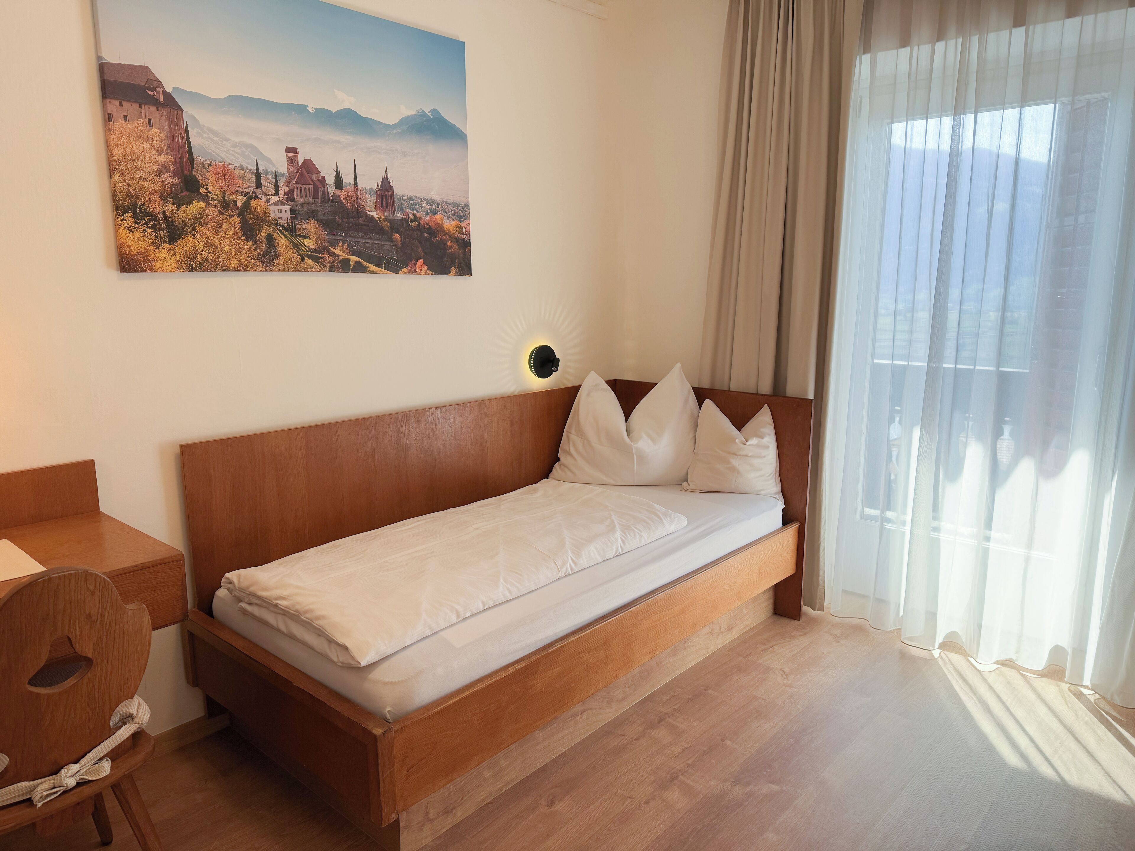 A single bed with a wooden headboard, a bedside table, a chair, and a painting of a mountainous landscape on the wall.