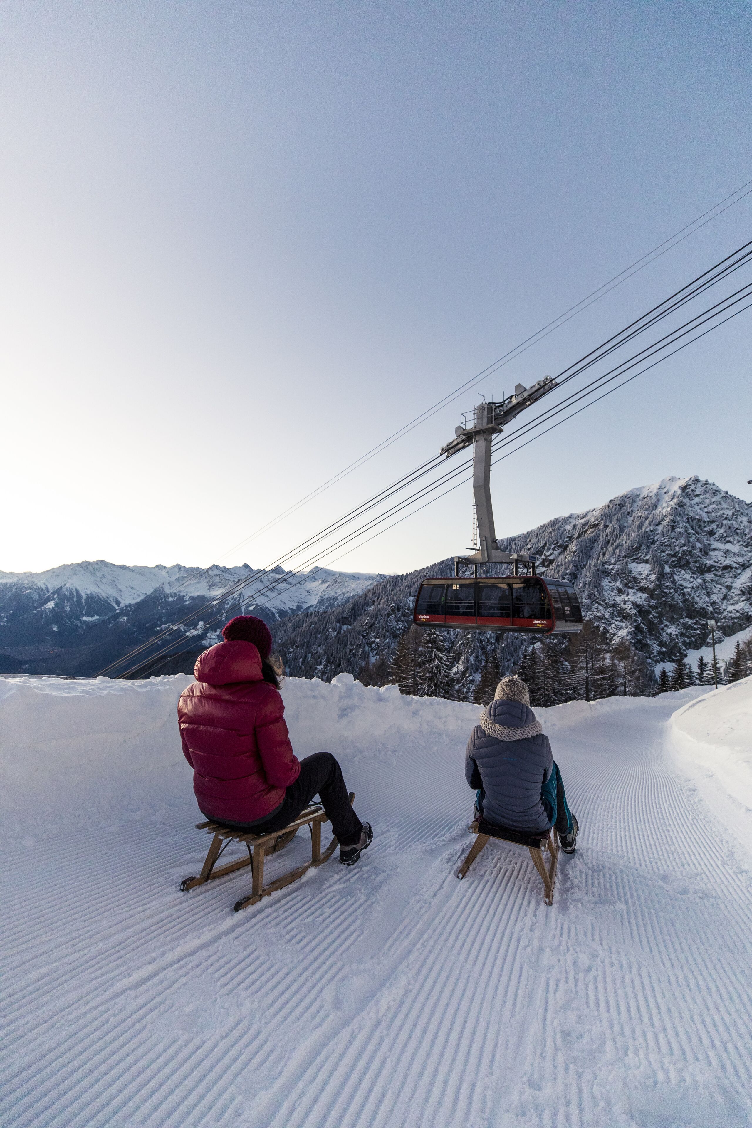 Two people sitting on wooden sleds on a snow-covered slope with a ski lift in the background.