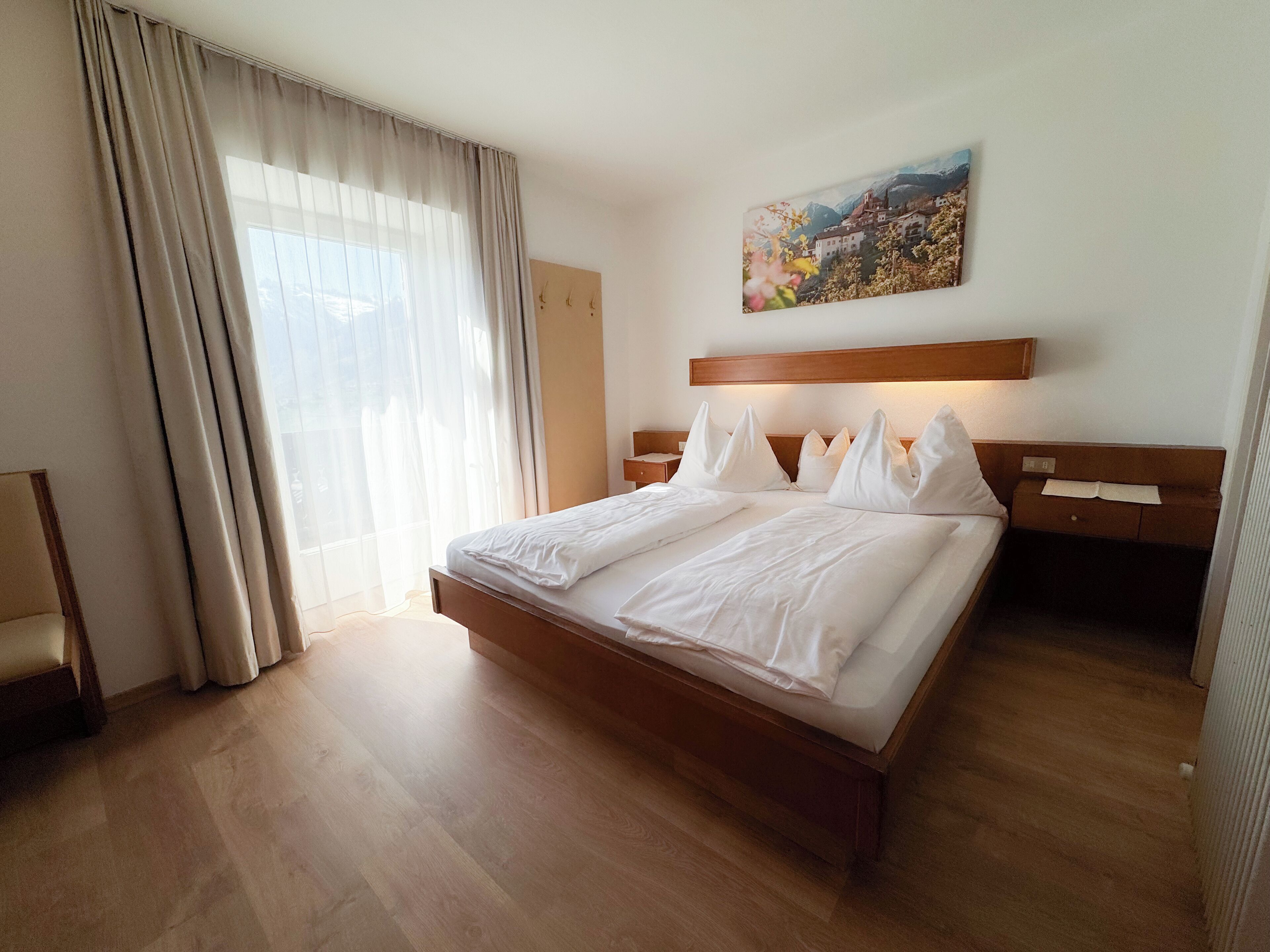 A hotel room with a large bed, wooden headboard, a nightstand, a bedside table, a window with sheer curtains, and a wall-mounted painting.