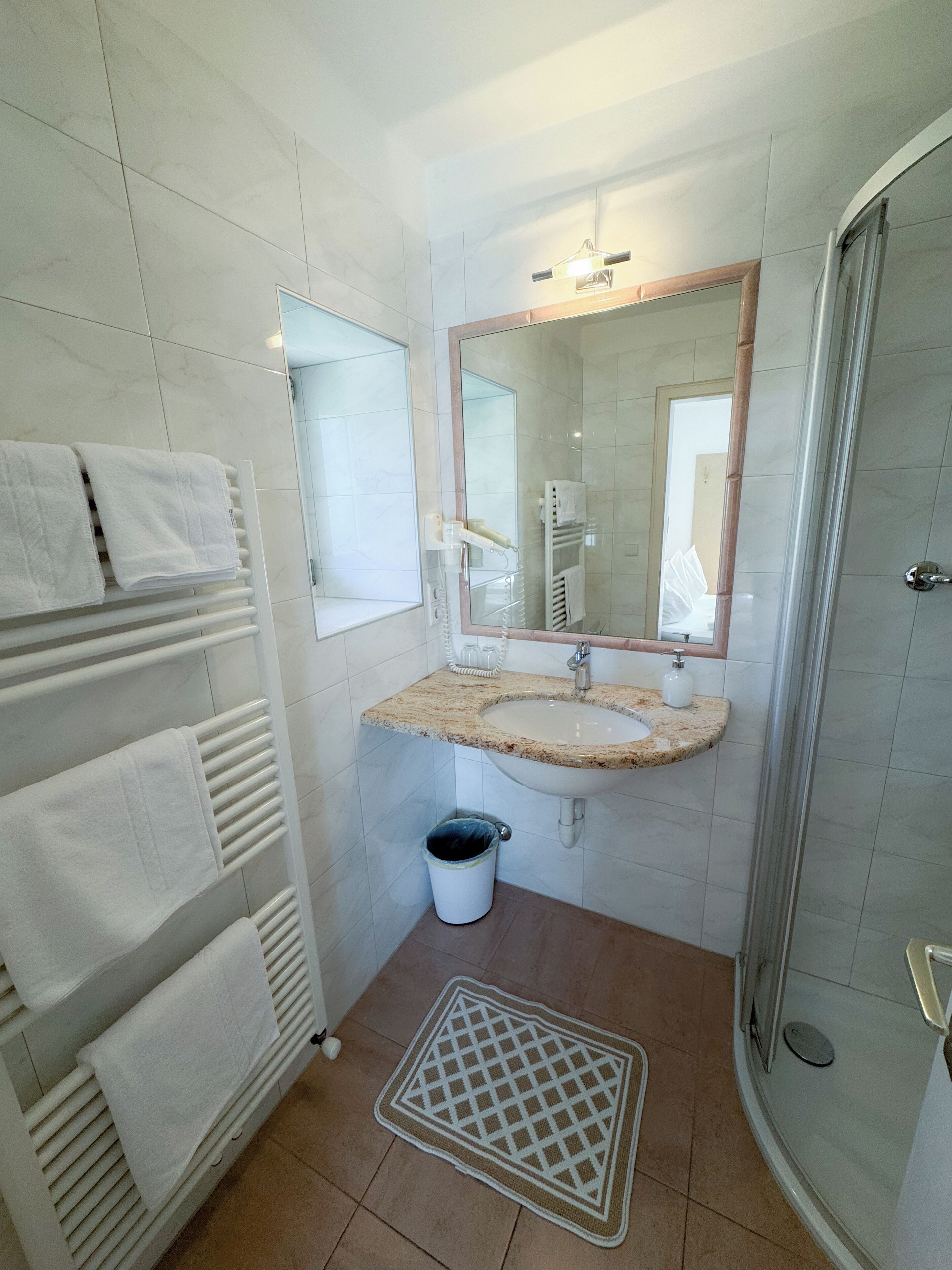 A bathroom with a white tiled floor, a sink with a marble countertop, a mirror above the sink, a towel rack with white towels, and a shower area with a glass door.