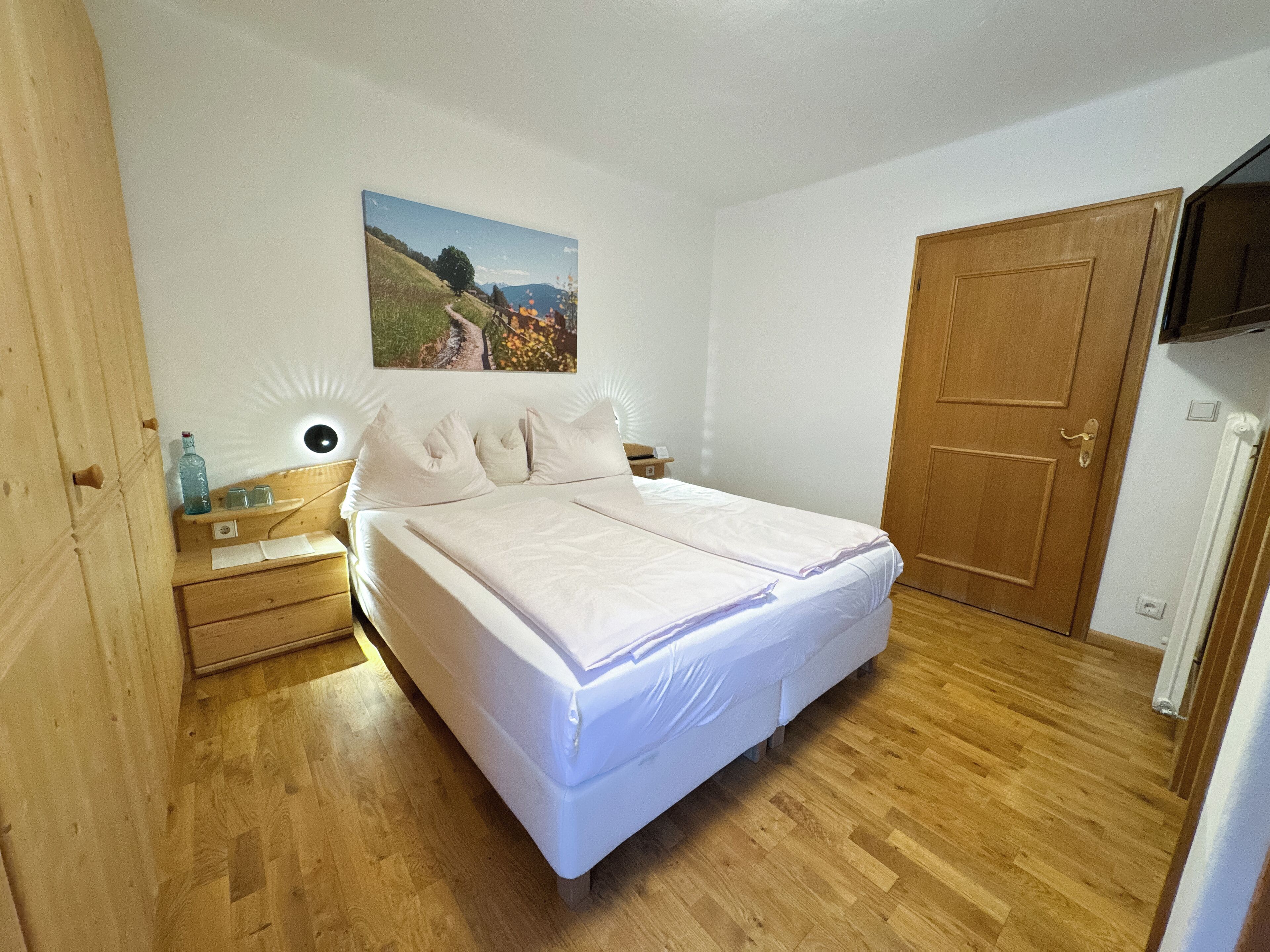 A bedroom with a wooden floor, a bed with white bedding, a nightstand with a lamp, a wooden door, and a wall-mounted TV.