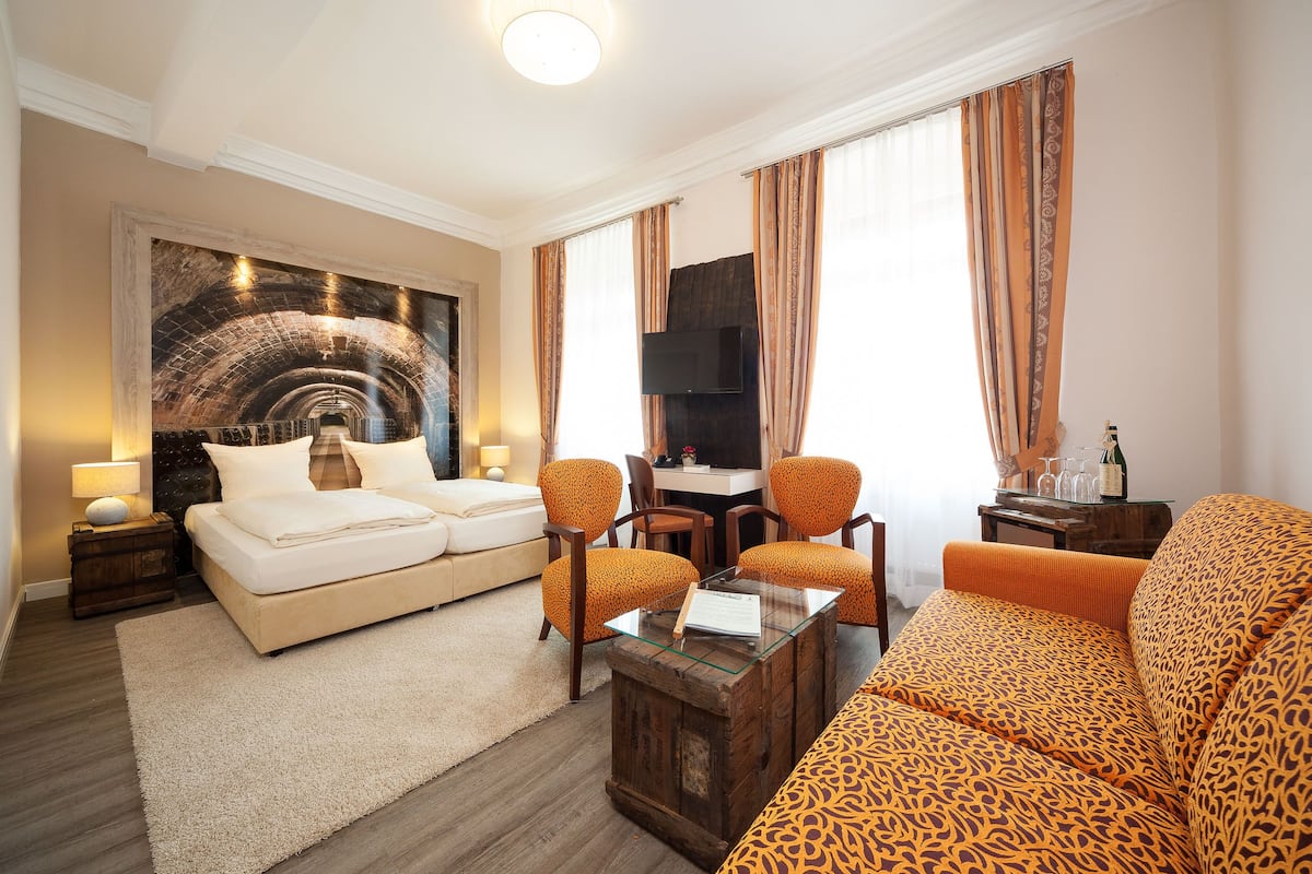 A hotel room with a bed, two armchairs, a sofa, a TV, and a large painting.