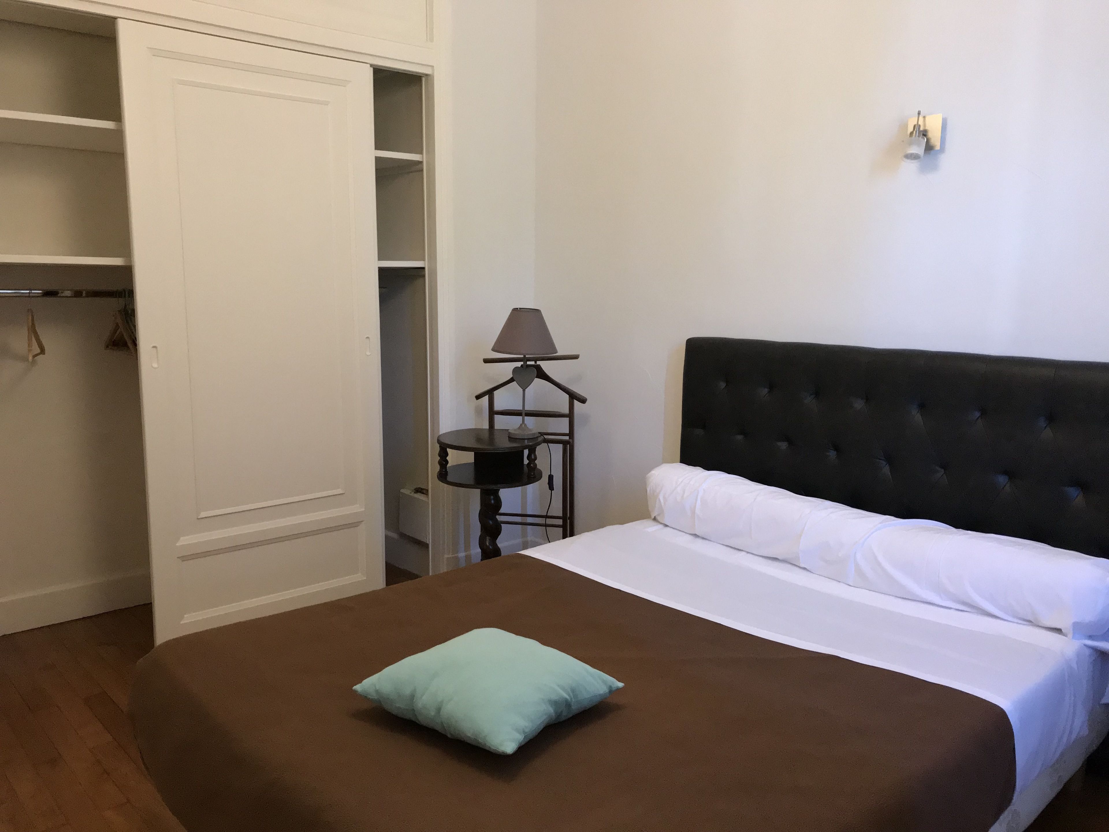 Un lit bien fait, recouvert d’une couverture marron et agrémenté d’un seul oreiller bleu clair, une tête de lit sombre en tissu, une table de chevet avec une lampe et une armoire blanche dont les portes sont ouvertes.