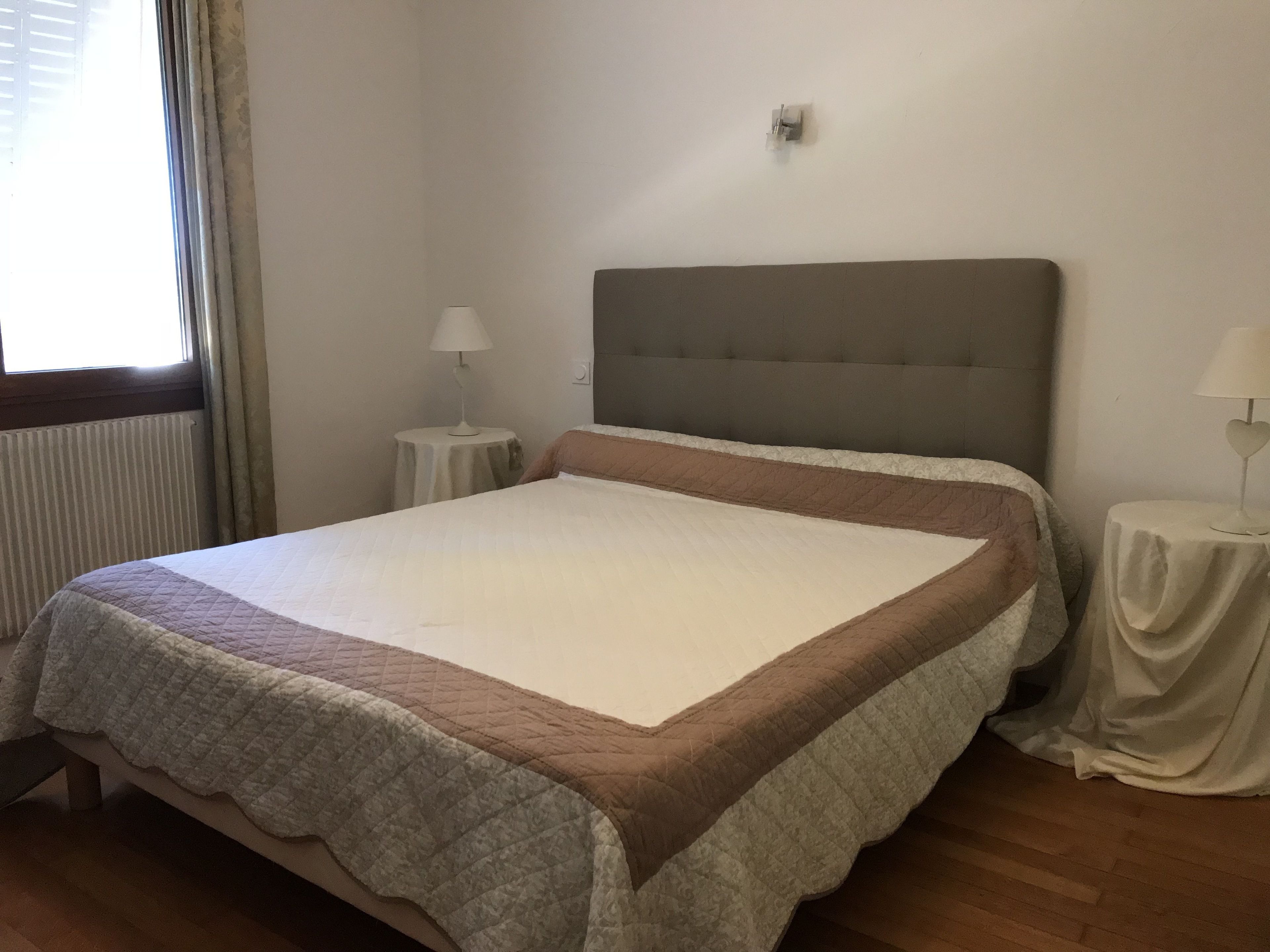 A neatly made bed with a quilted bedspread and a padded headboard in a room with a radiator, two bedside tables with lamps, and a window with curtains.