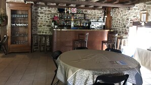 Un coin bar rustique avec des murs en pierre, des poutres en bois et un comptoir où sont exposées diverses bouteilles et verres. Des chaises sont disposées autour d’une table recouverte d’une nappe.
