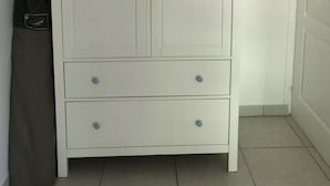 Une armoire blanche, dotée de deux tiroirs et d’une porte, est placée dans un coin. Un sac gris est accroché au mur, à sa gauche.