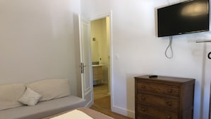 Une chambre à coucher comprenant un lit, une commode en bois, un téléviseur fixé au mur et une porte donnant sur une salle de bain.