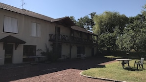 Un bâtiment doté d’un toit en tuiles, d’un balcon orné d’un motif en treillis et d’un espace verdoyant aménagé avec des sièges d’extérieur.