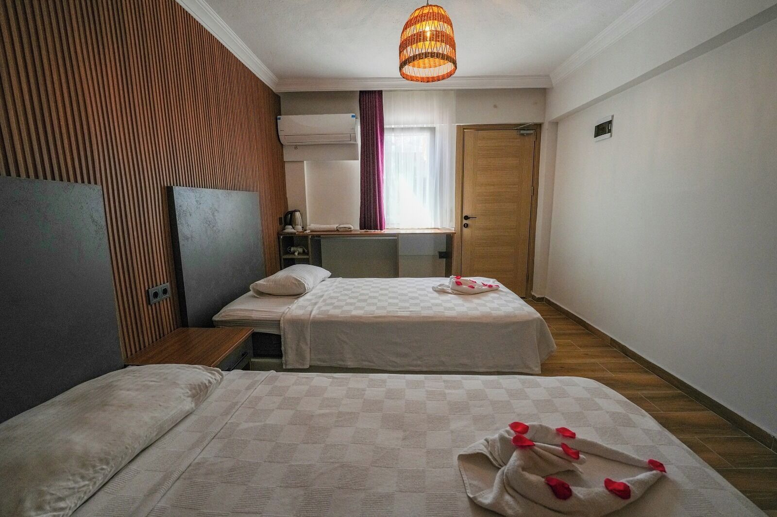 A hotel room with two beds, a wooden headboard, a large window with curtains, a wooden door, and a hanging light fixture.