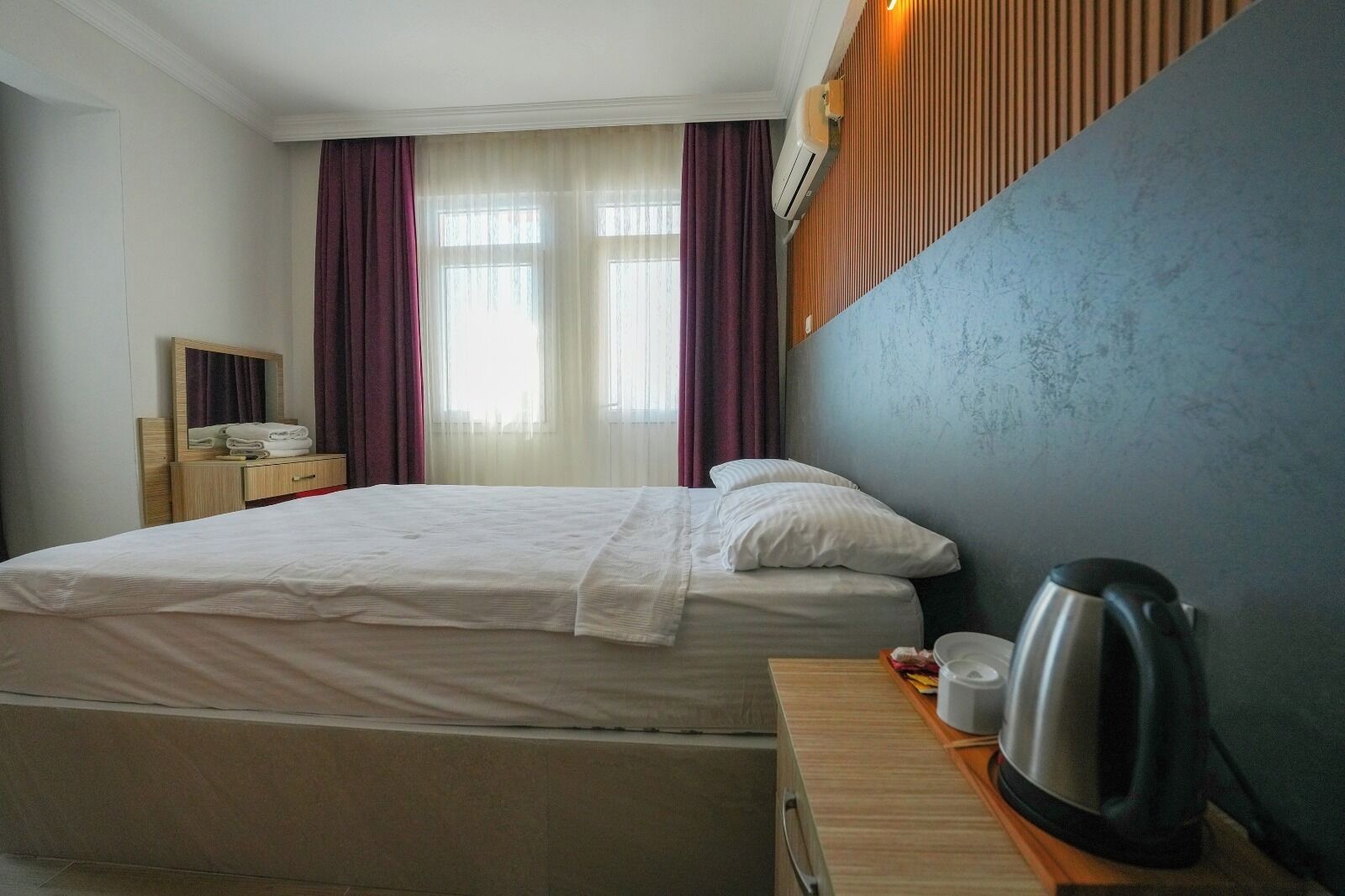 Habitación de hotel con cama, mesita de noche con hervidor, espejo y ventana con cortinas.